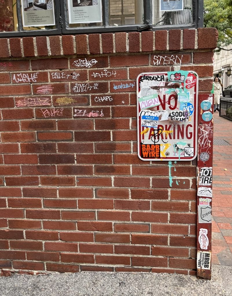 graffiti and stickers on the side of a brick building with a "no parking" sign