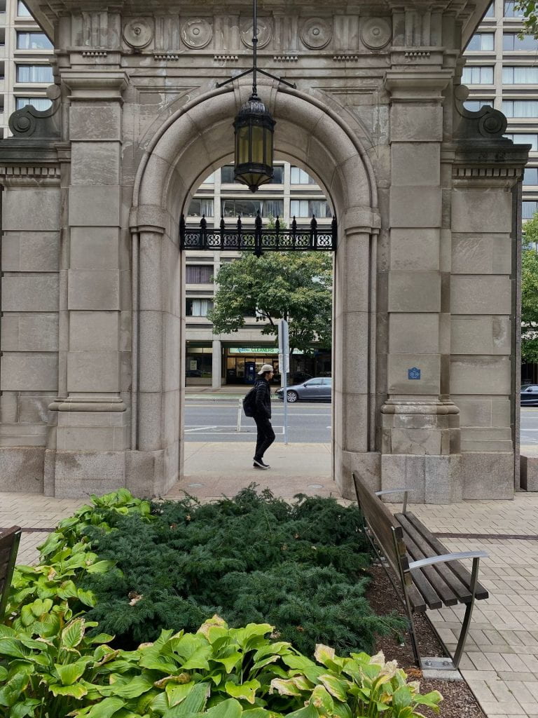 person walking through an ornate concrete archway with greenery in the foreground